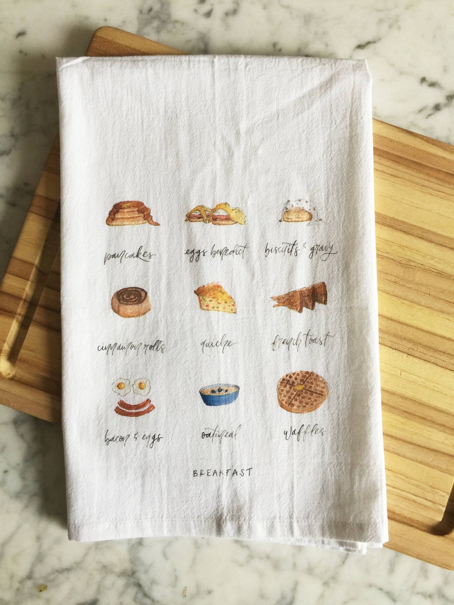 A white tea towel displays 9 breakfast foods: pancakes, eggs benedict, biscuits and gravy, cinnamon roll, quiche, french toast, bacon and eggs, oatmeal, and waffles
