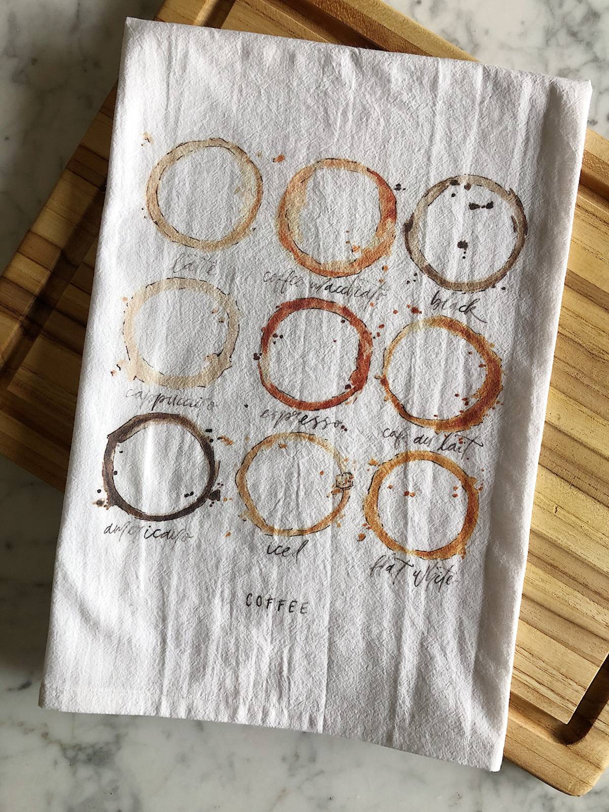 A white tea towel displaying watercolor illustrations of 9 different types of coffees as coffee ring stains