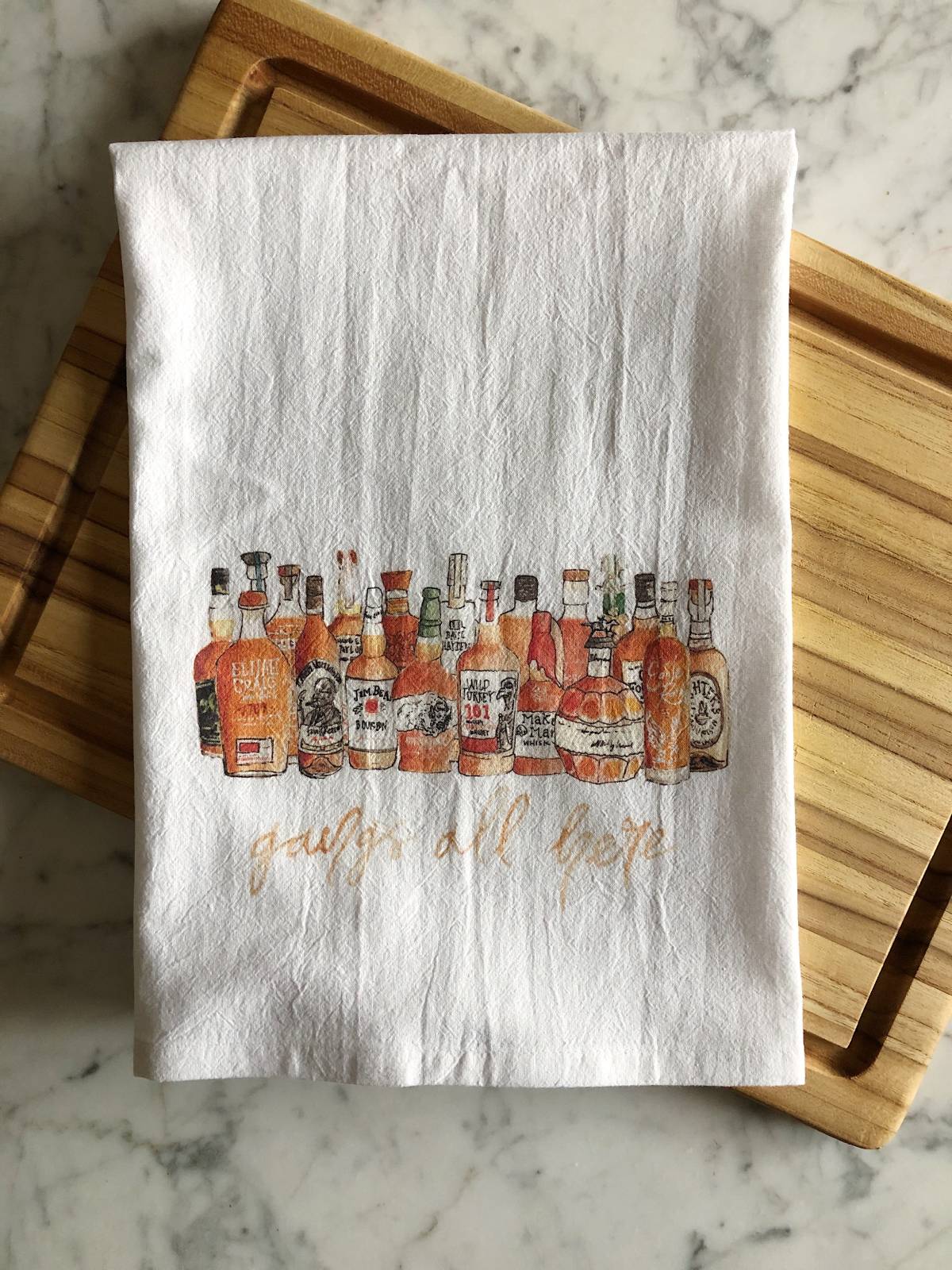 A white tea towel displaying watercolor illustrations of multiple popular bourbons with the phrase "gangs all here" beneath