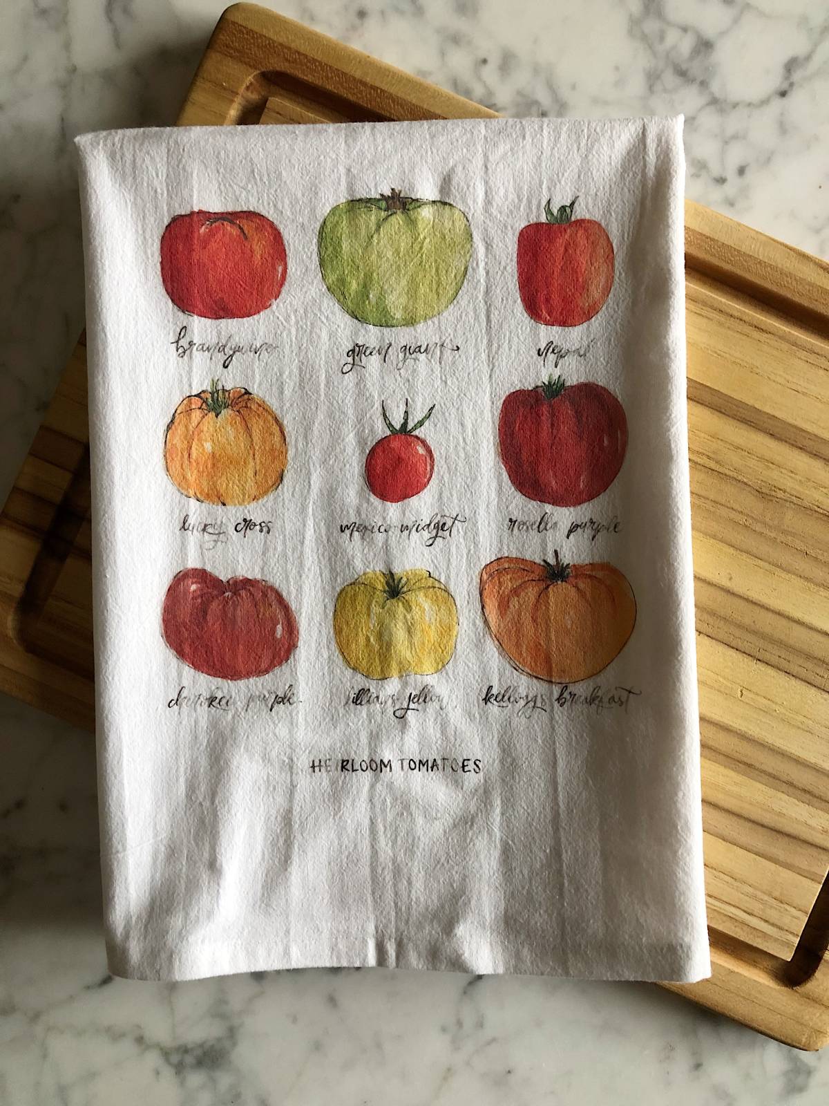 A white tea towel displaying watercolor illustrations of 9 heirloom tomatoes