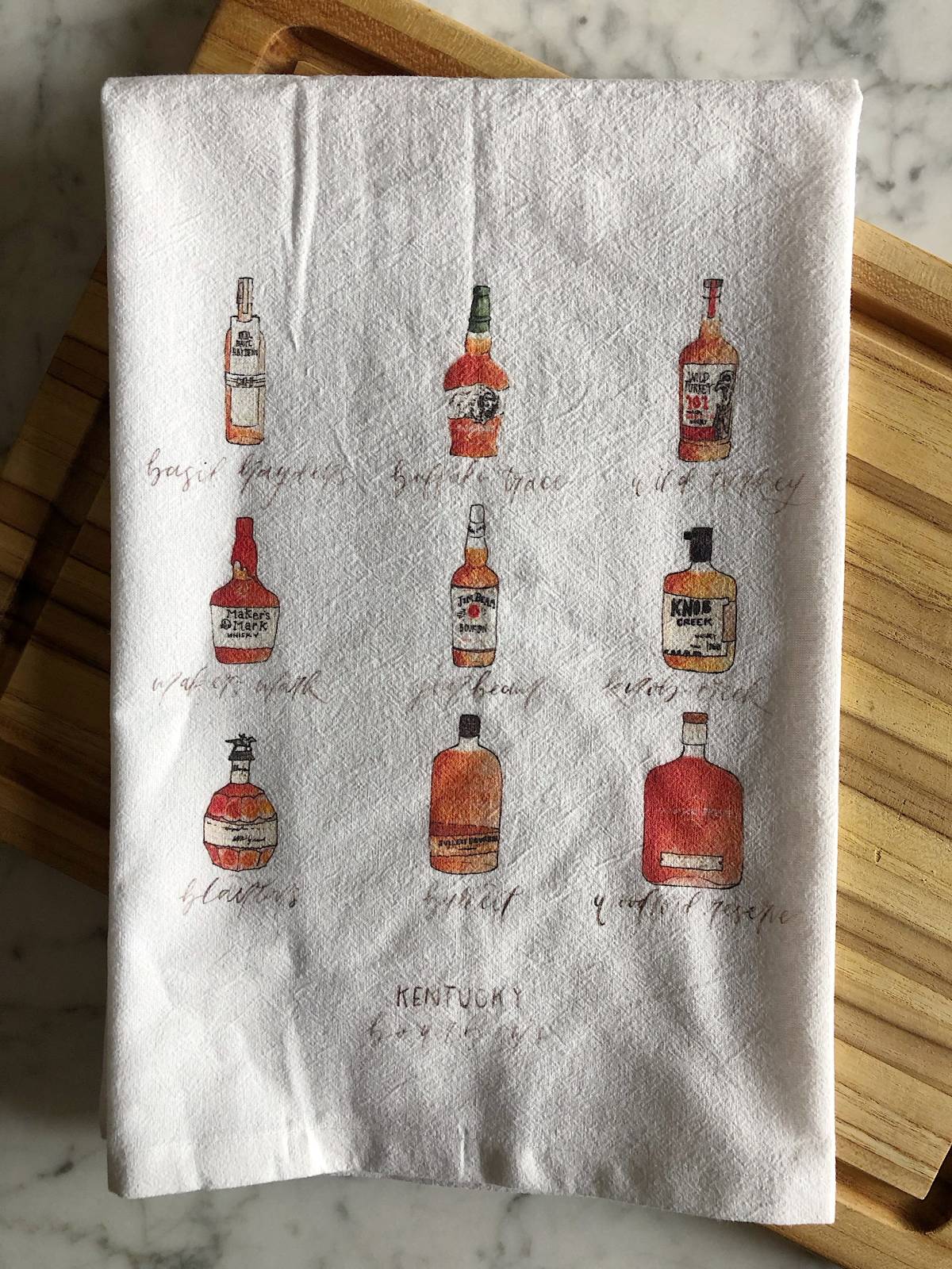 A white tea towel displaying watercolor illustrations of 9 popular Kentucky bourbons