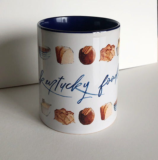 A white ceramic mug displays watercolor illustrations of 9 popular Kentucky foods and drinks. In the middle is the phrase "kentucky foodie"